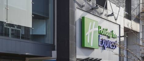 A Holiday Inn Express building with a glass facade, a visible entrance, and a fire alarm box.