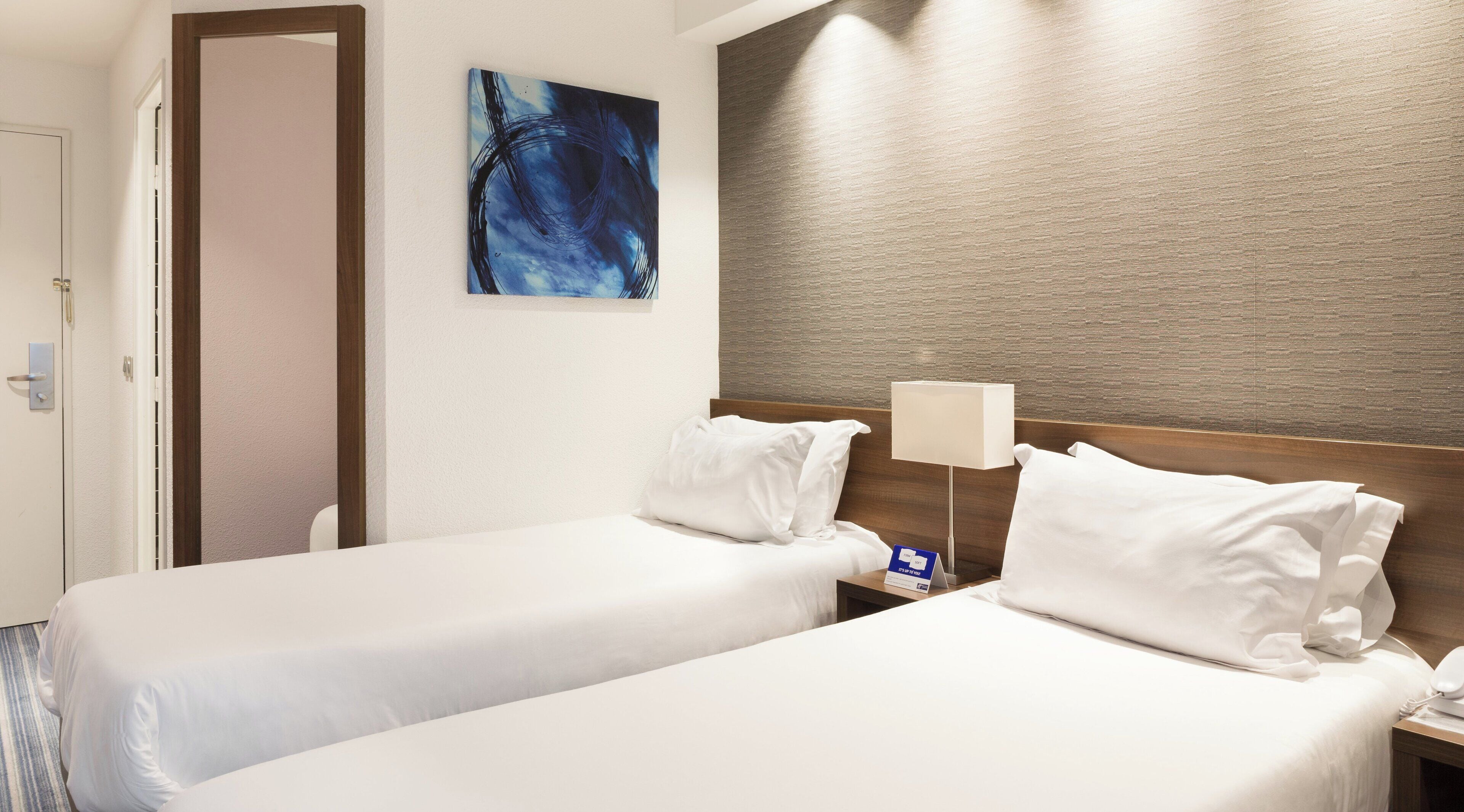 A hotel room with two beds, a painting on the wall, and a textured wall panel.