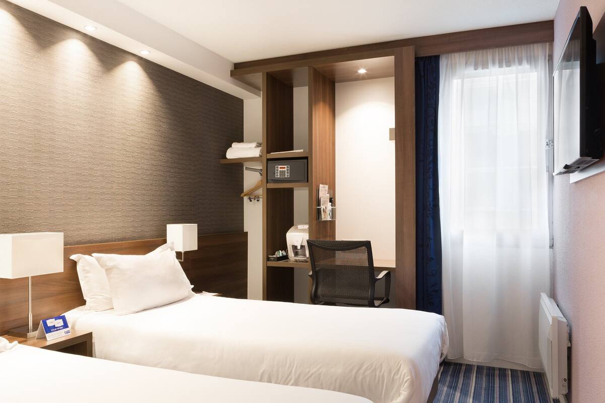 A hotel room with two beds, a desk, a chair, a TV, and a window with curtains.