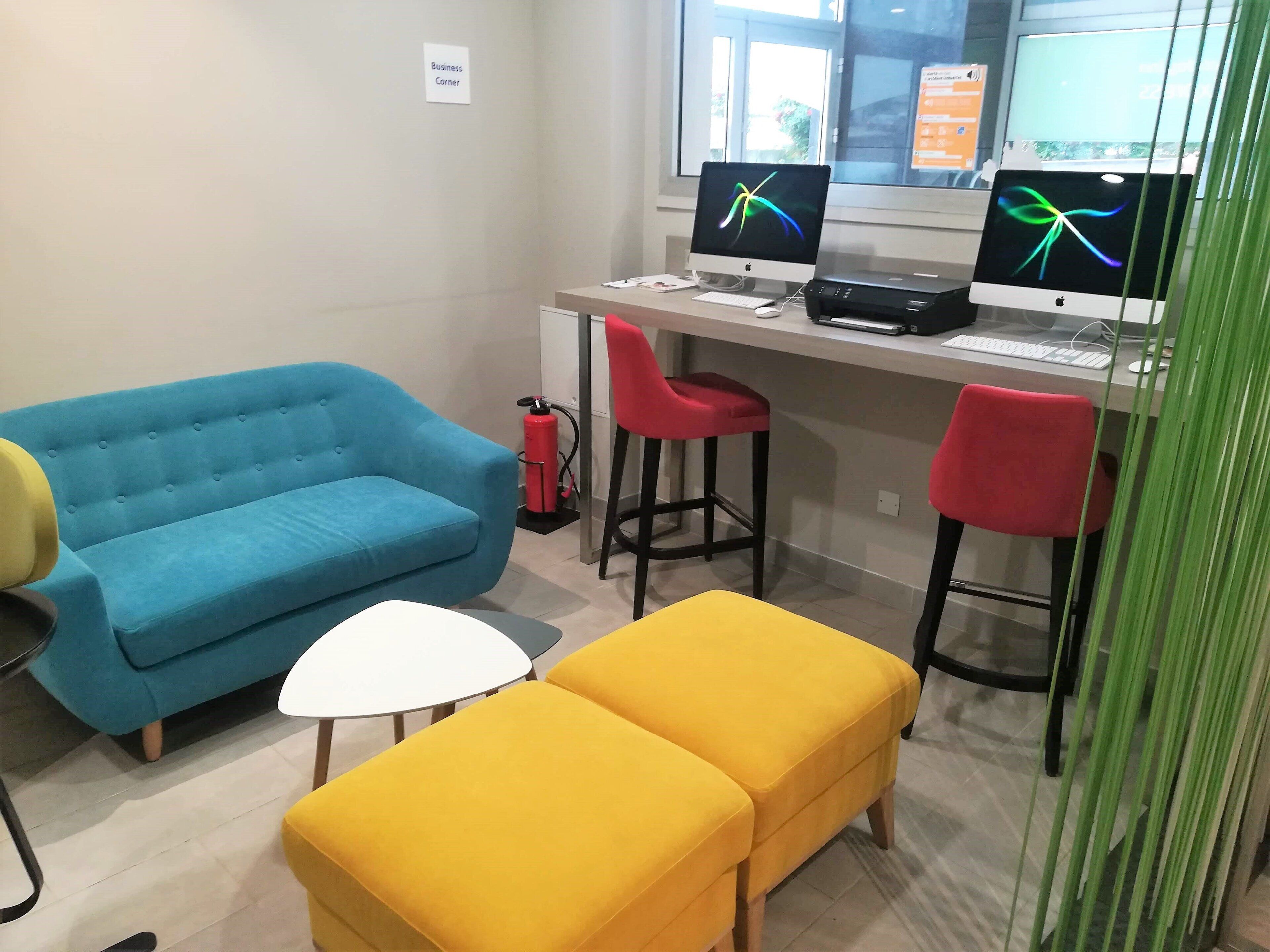 A workspace with a desk, two monitors, a blue sofa, a yellow ottoman, and red chairs.