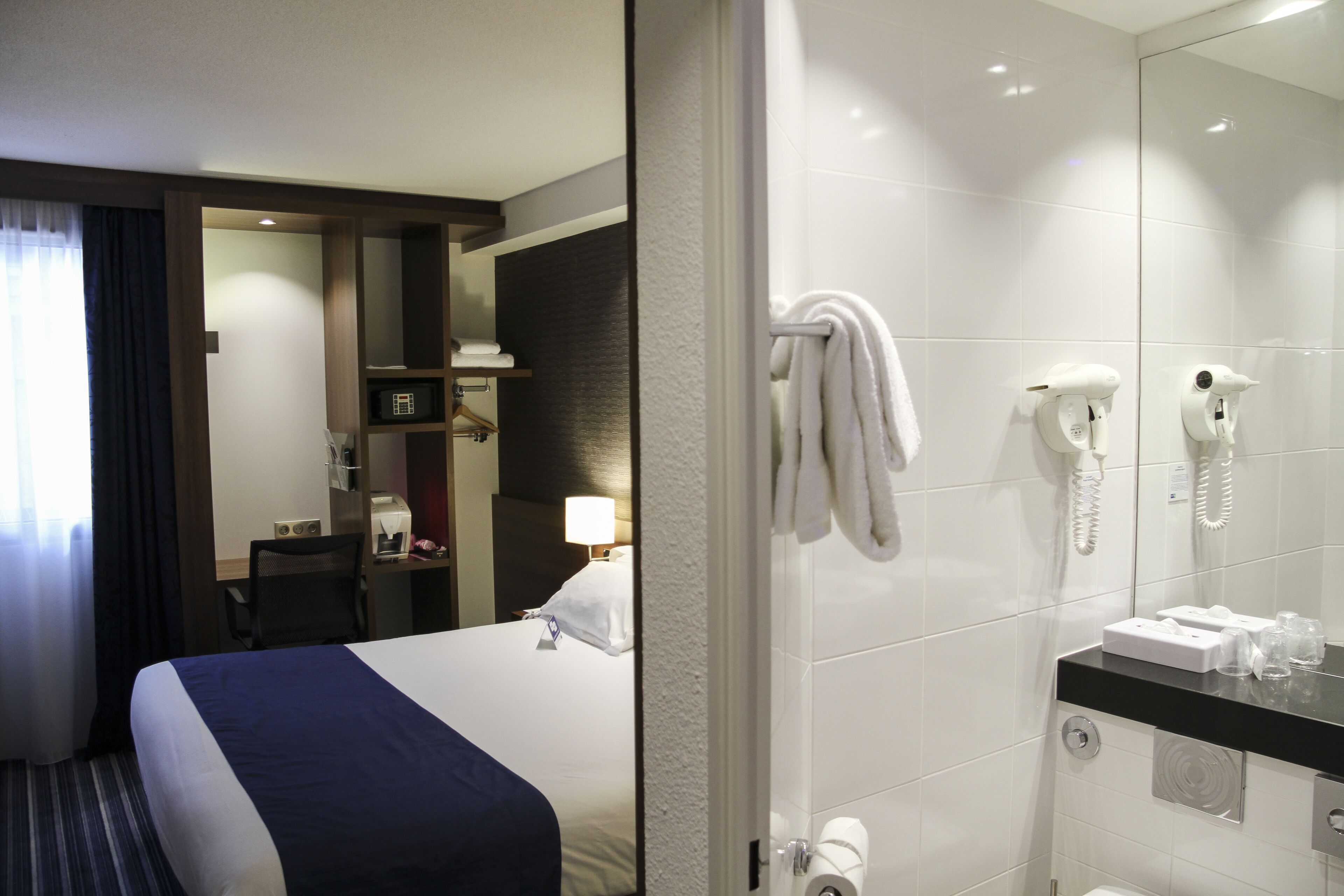 A hotel room with a bed, a desk, a chair, a mirror, a bathroom with a sink, a toilet, and a shower.