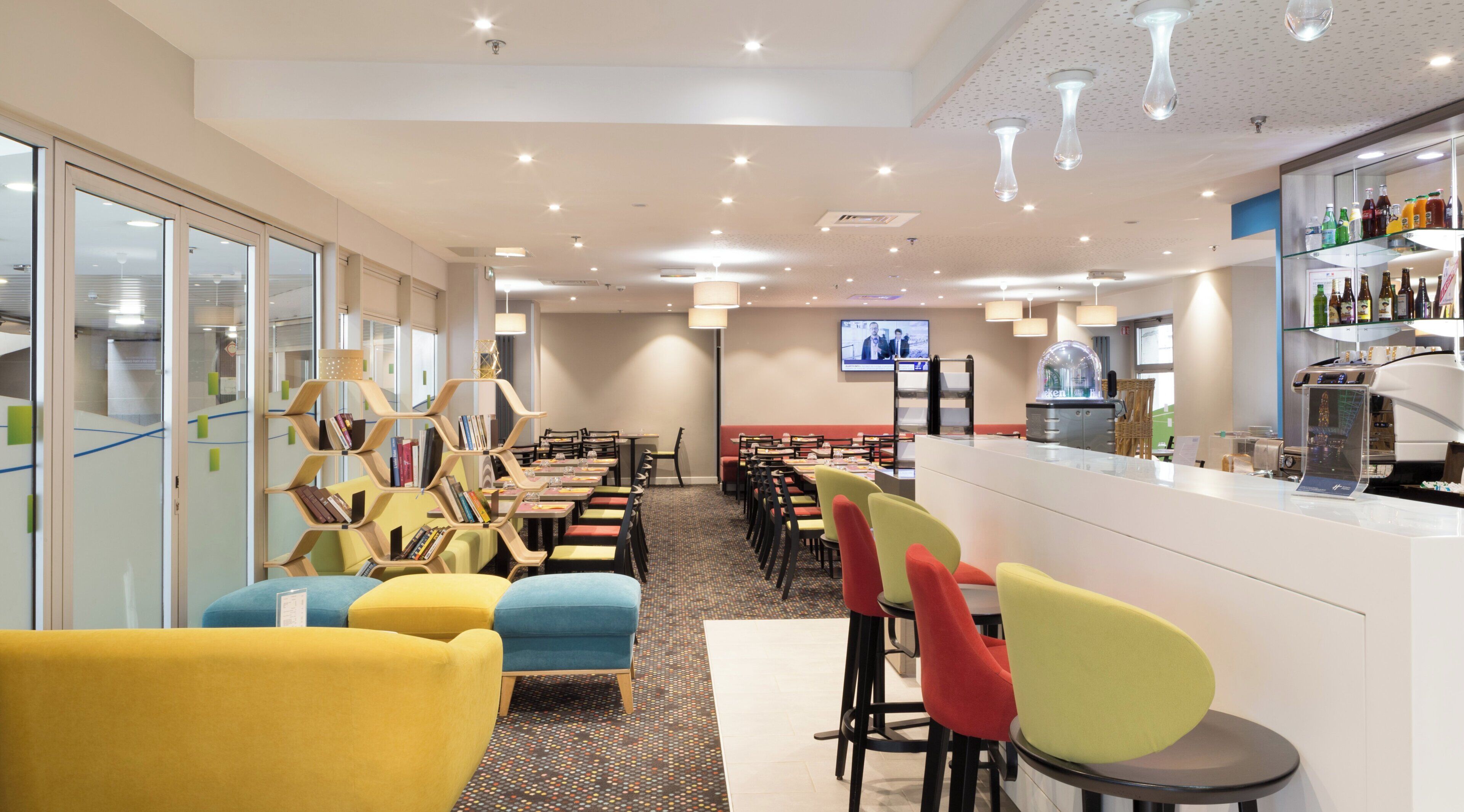 A hotel dining area with multiple tables and chairs, a bar, and a television screen.
