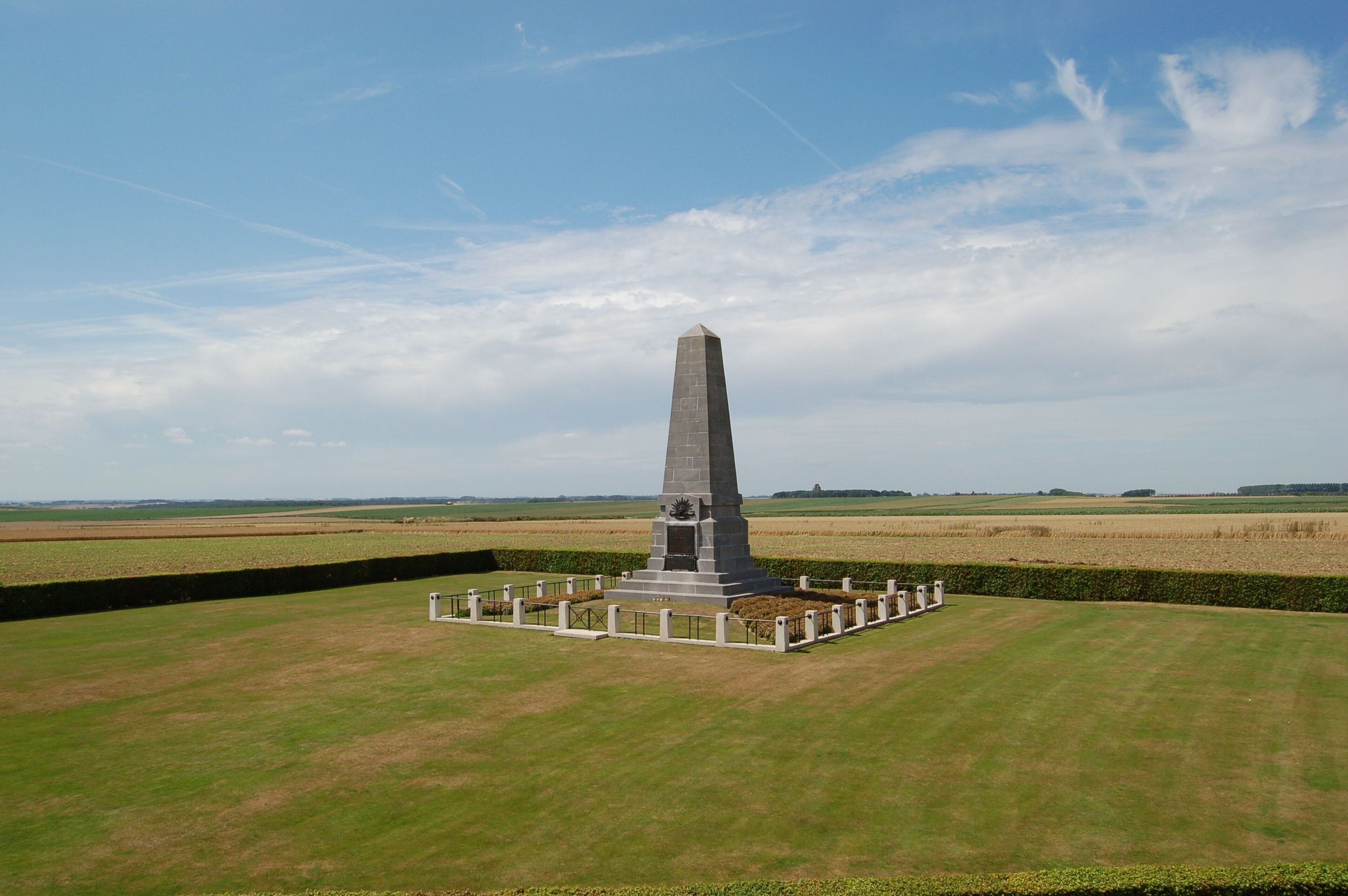 Un monument haut et étroit, doté d’une base entourée d’une petite clôture, se dresse au milieu d’un champ verdoyant, avec en arrière-plan d’autres champs et un ciel dégagé.