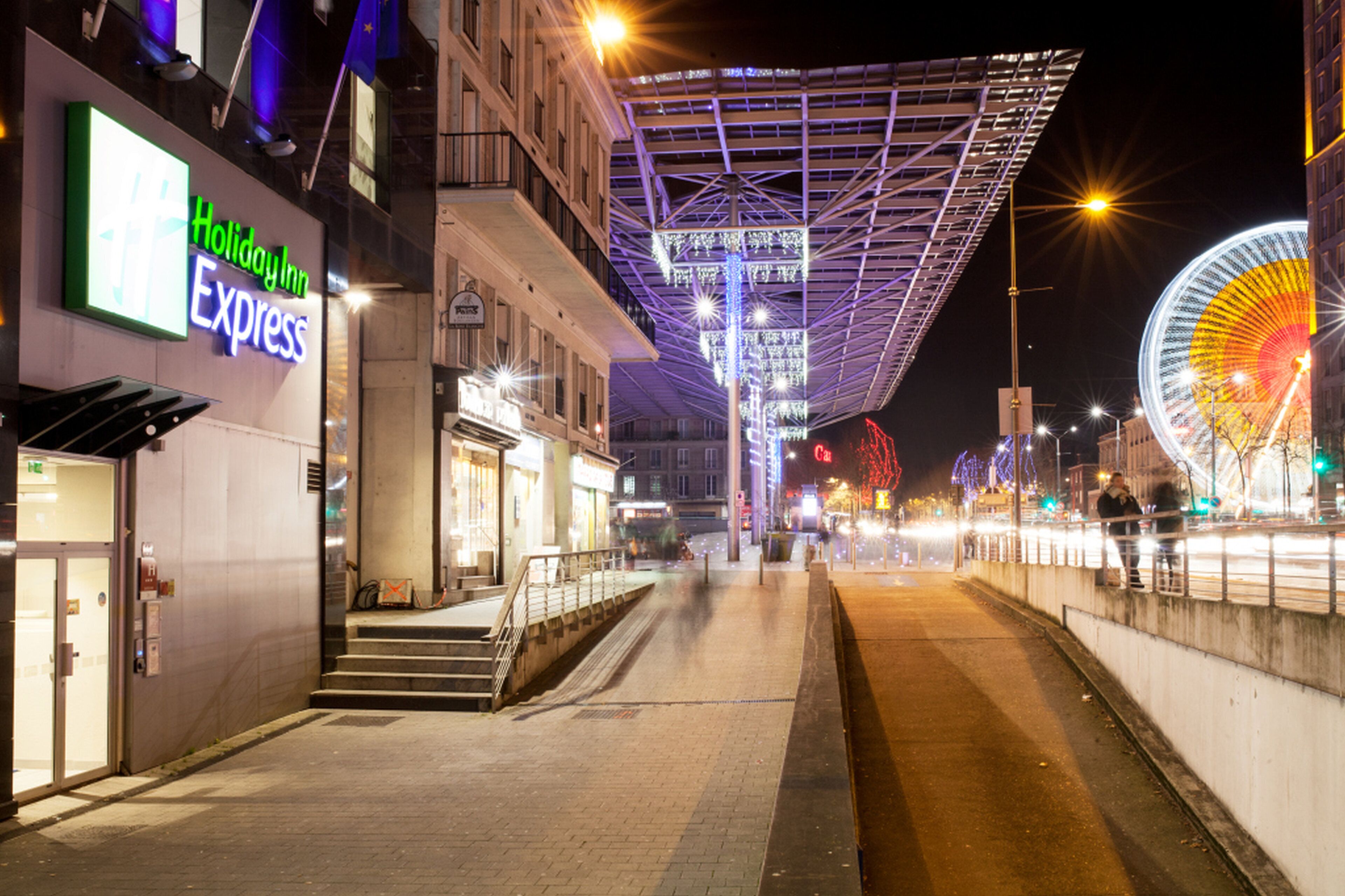 Ein nächtlicher Straßenblick mit einem Holiday Inn Express Schild, einem Riesenrad und einem modernen Gebäude mit einer markanten Dachkonstruktion.