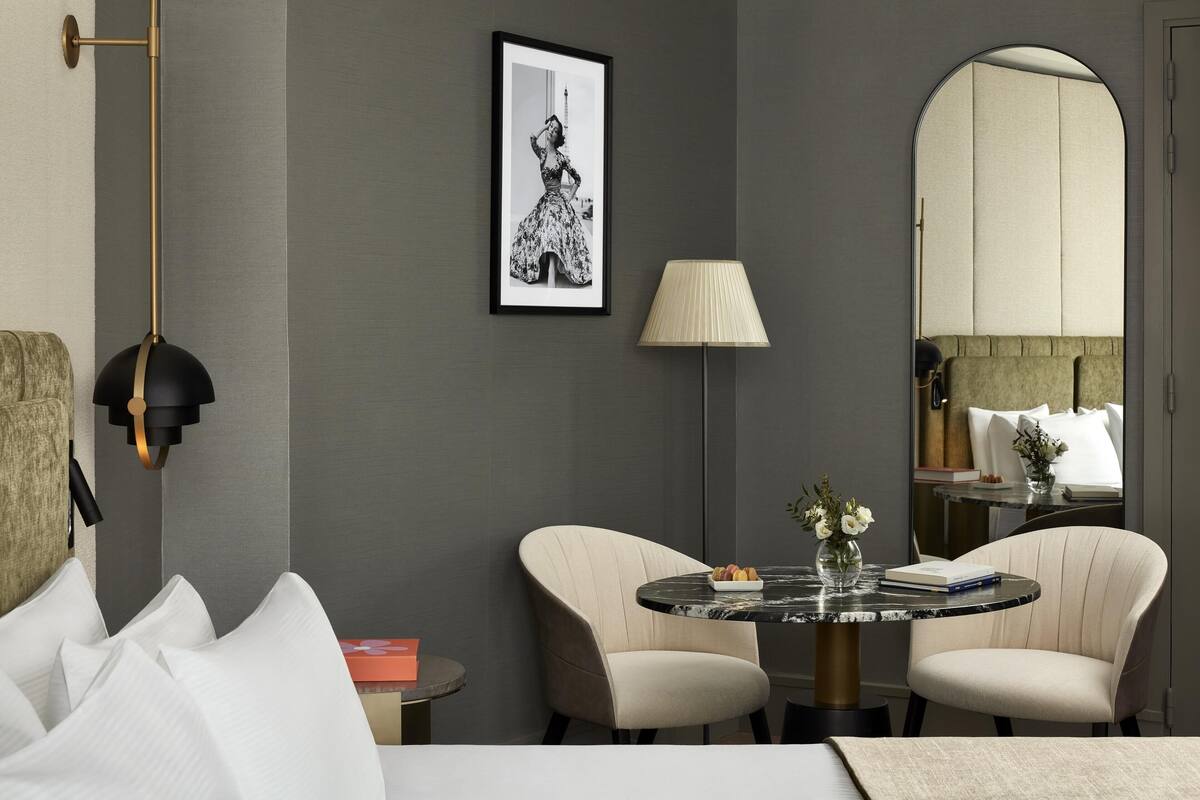 A modern hotel room with a round marble table, two upholstered chairs, a large mirror, a framed picture on the wall, and a floor lamp.