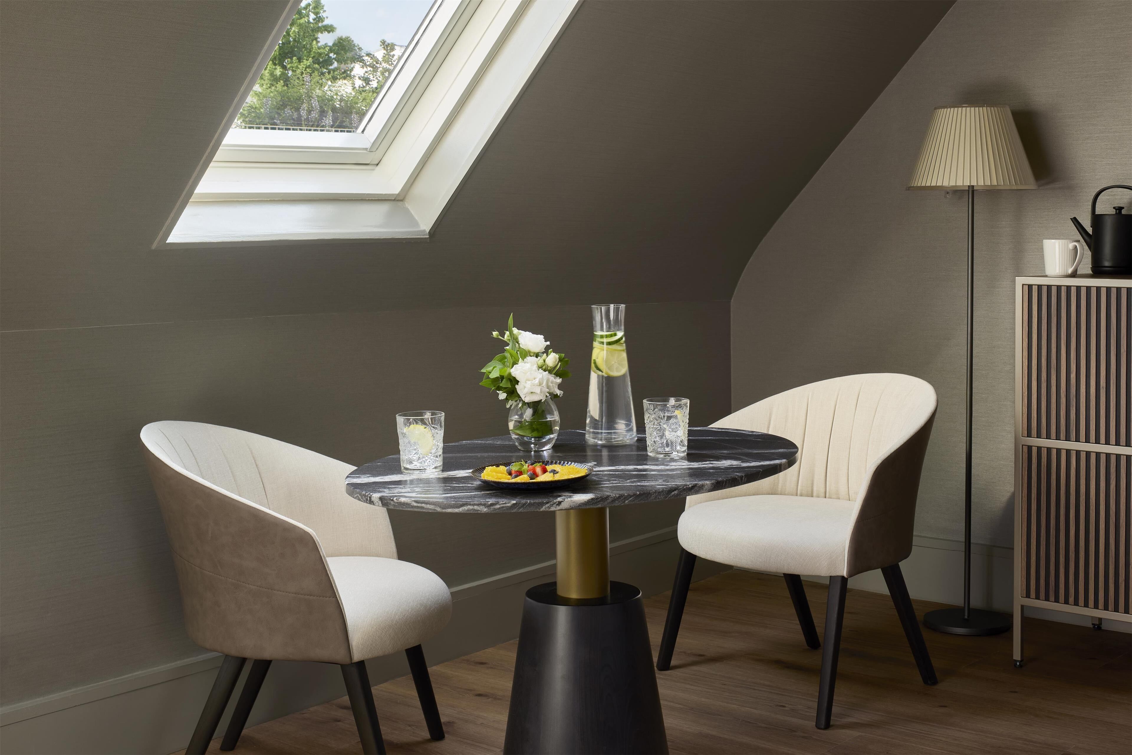 A modern dining area with a round table, two chairs, a vase with flowers, a glass of water, and a lamp.