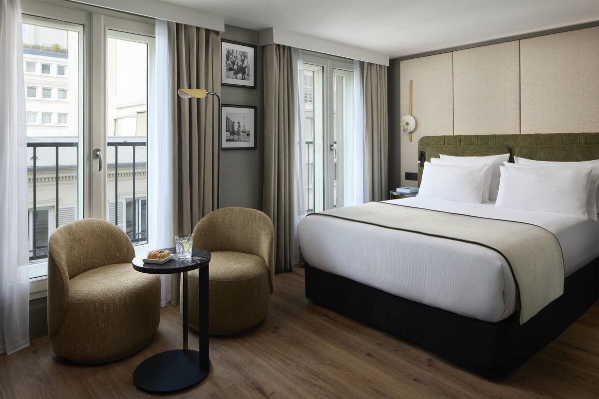 A hotel room with a large bed, two armchairs, a small table with a tray of pastries, and a view of the city through large windows.