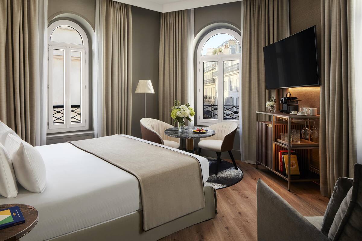 A hotel room with a bed, a desk, a chair, a TV, and a bookshelf.