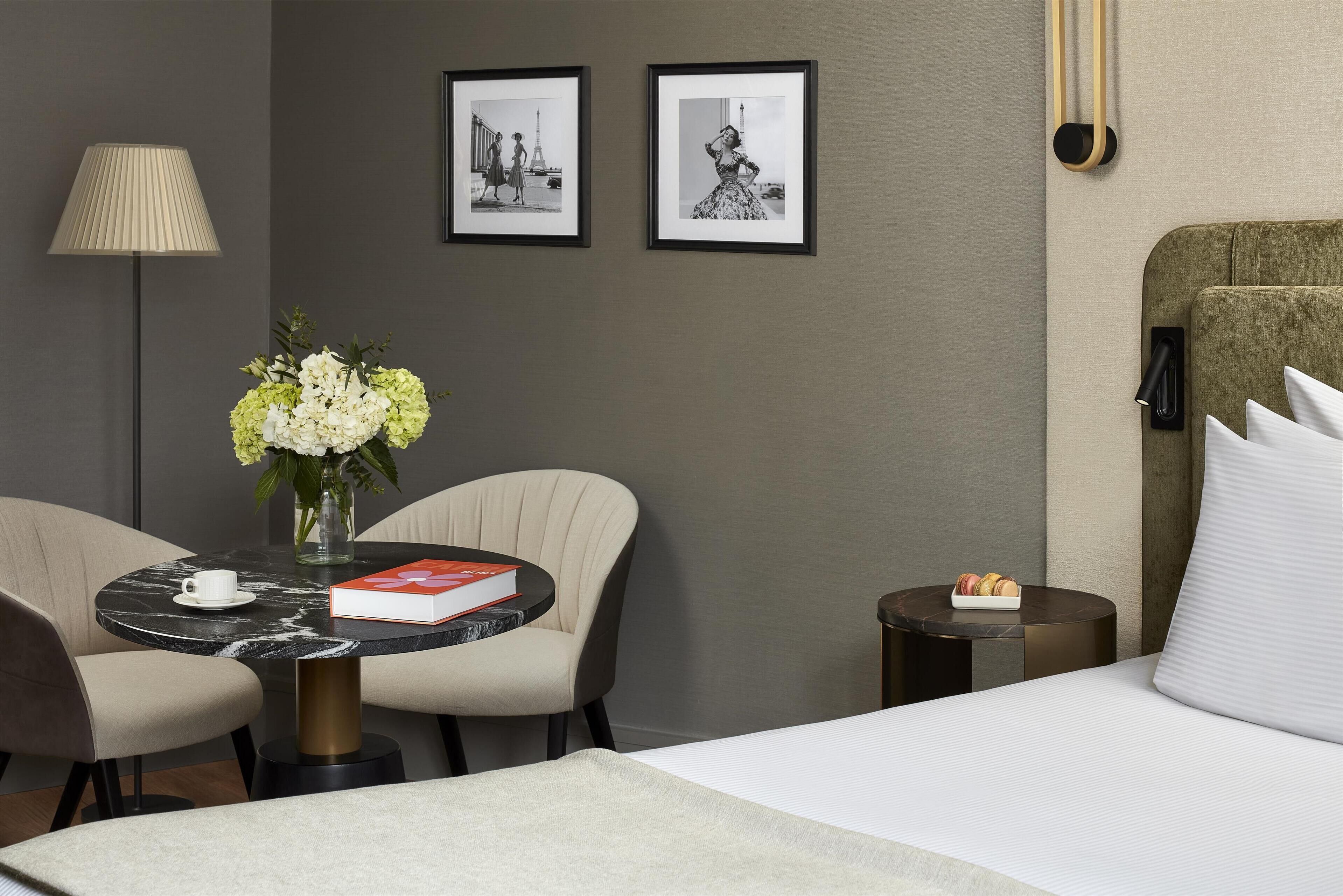 Habitación de hotel con cama, una mesa redonda con sillas, un florero, una lámpara y dos cuadros en la pared.