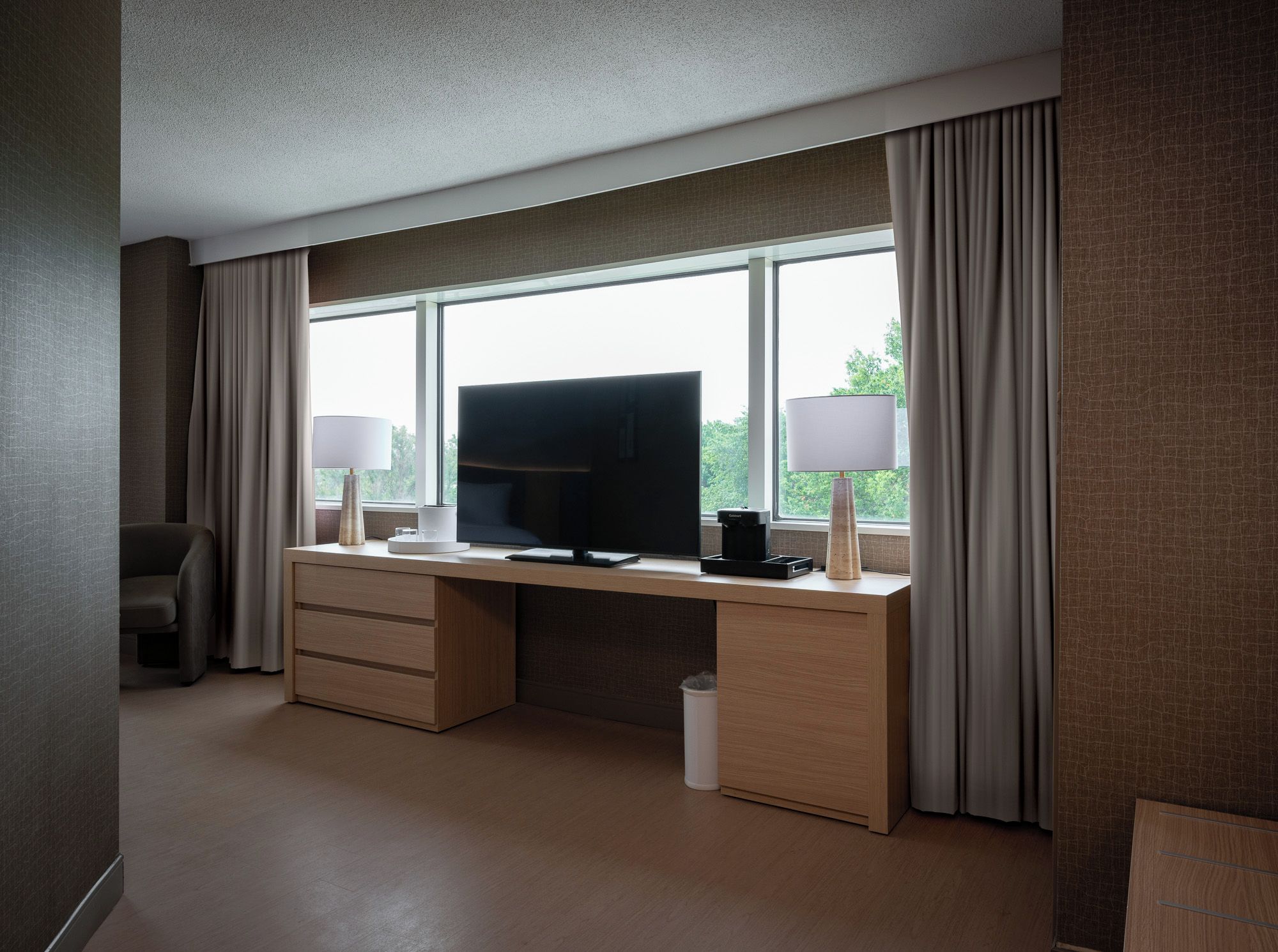 A hotel room with a large window, a flat-screen TV, a desk with two lamps, and a chair.