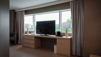 A hotel room with a large window, a flat-screen TV, a desk with two lamps, and a chair.