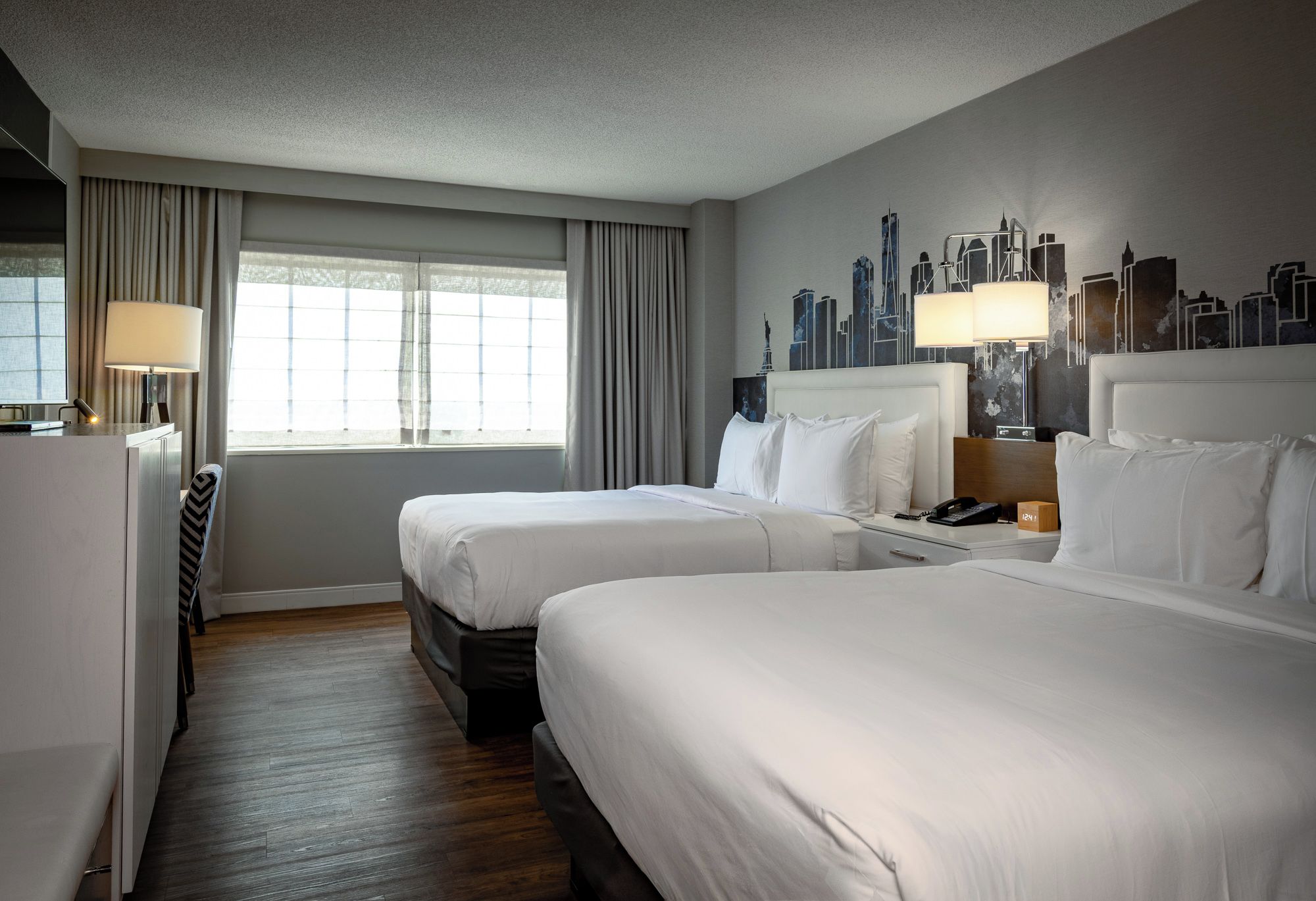 A hotel room with two beds, a desk, a chair, a lamp, and a cityscape mural on the wall.