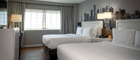 A hotel room with two beds, a desk, a chair, a lamp, and a cityscape mural on the wall.