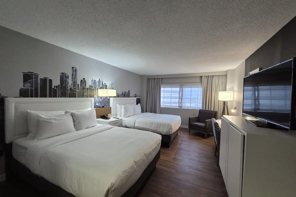 Ein Hotelzimmer mit zwei Betten, einem Fernseher, einem Schreibtisch und einem Wandgemälde mit Stadtansicht.