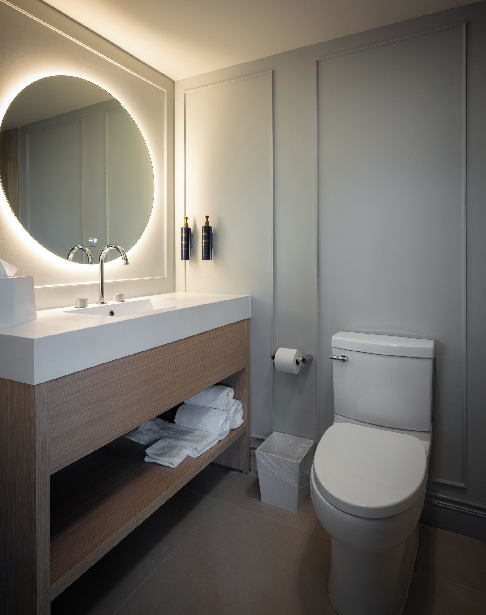 A modern bathroom with a round mirror, a white sink, a toilet, and a shelf with towels.