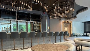 A modern bar area with multiple seating options, including round tables and cushioned chairs, and a bar counter with a view of the outdoors. - World of Blue (East Rutherford)