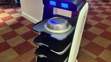 A self-service coffee machine with a digital display, located in a public area with checkered flooring and red chairs.