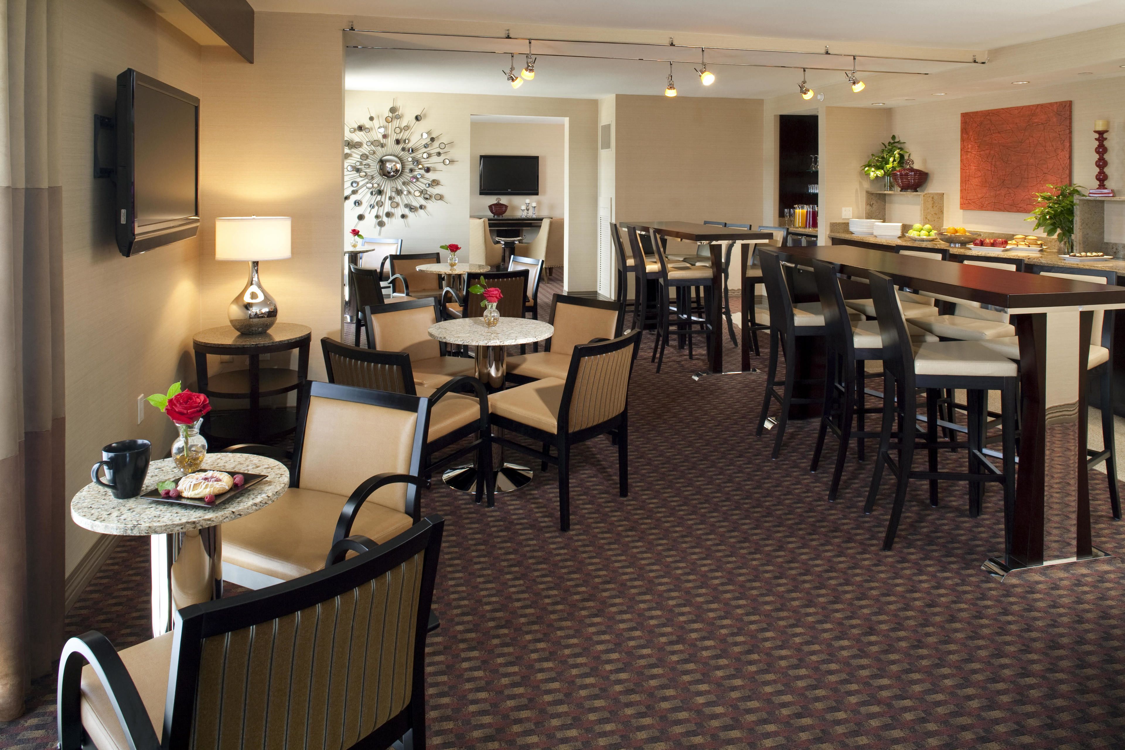 A hotel dining area with round tables, chairs, a TV, and a kitchen area with a fruit display.
