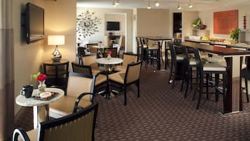 A hotel dining area with round tables, chairs, a TV, and a kitchen area with a fruit display.