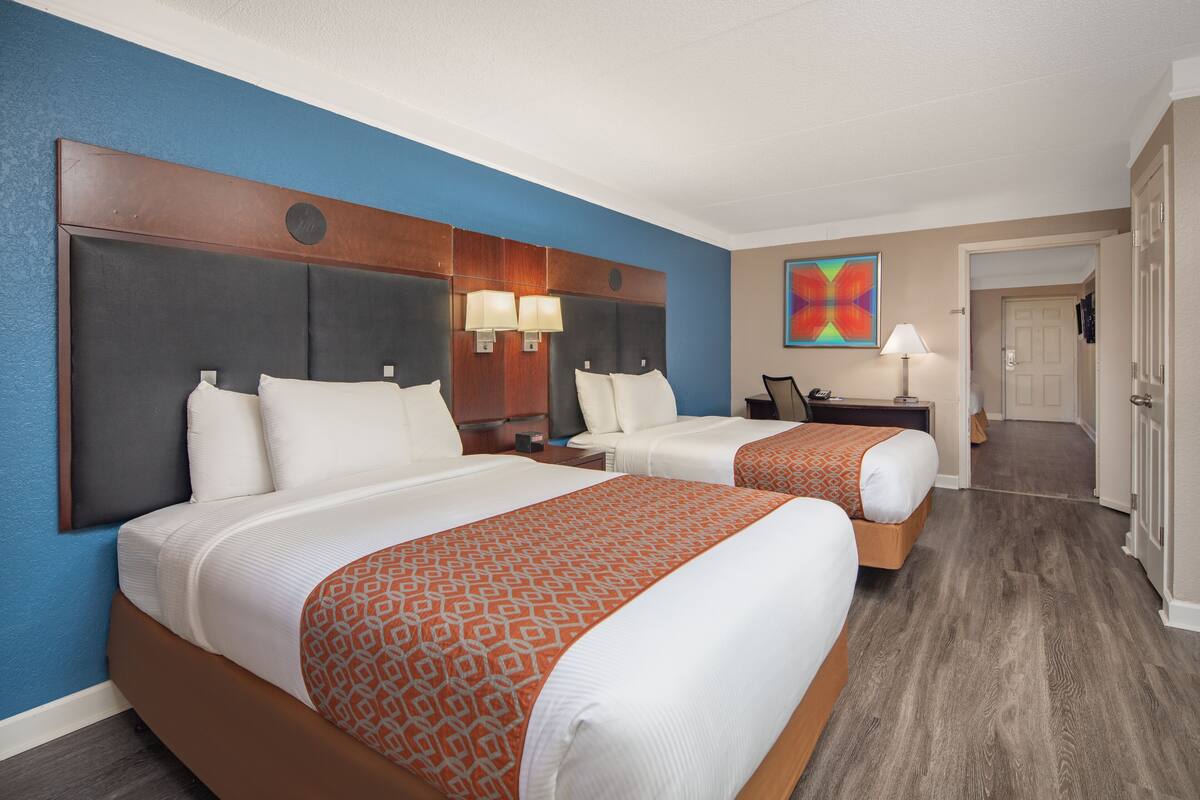 Habitación de hotel con dos camas, cabecera con lámparas integradas, un cuadro en la pared y una puerta que da a otra habitación.