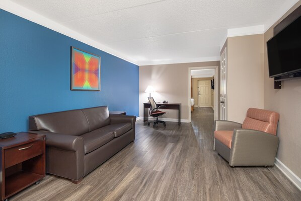 A hotel room with a brown leather sofa, a desk with a lamp, a TV mounted on the wall, and a wooden floor.