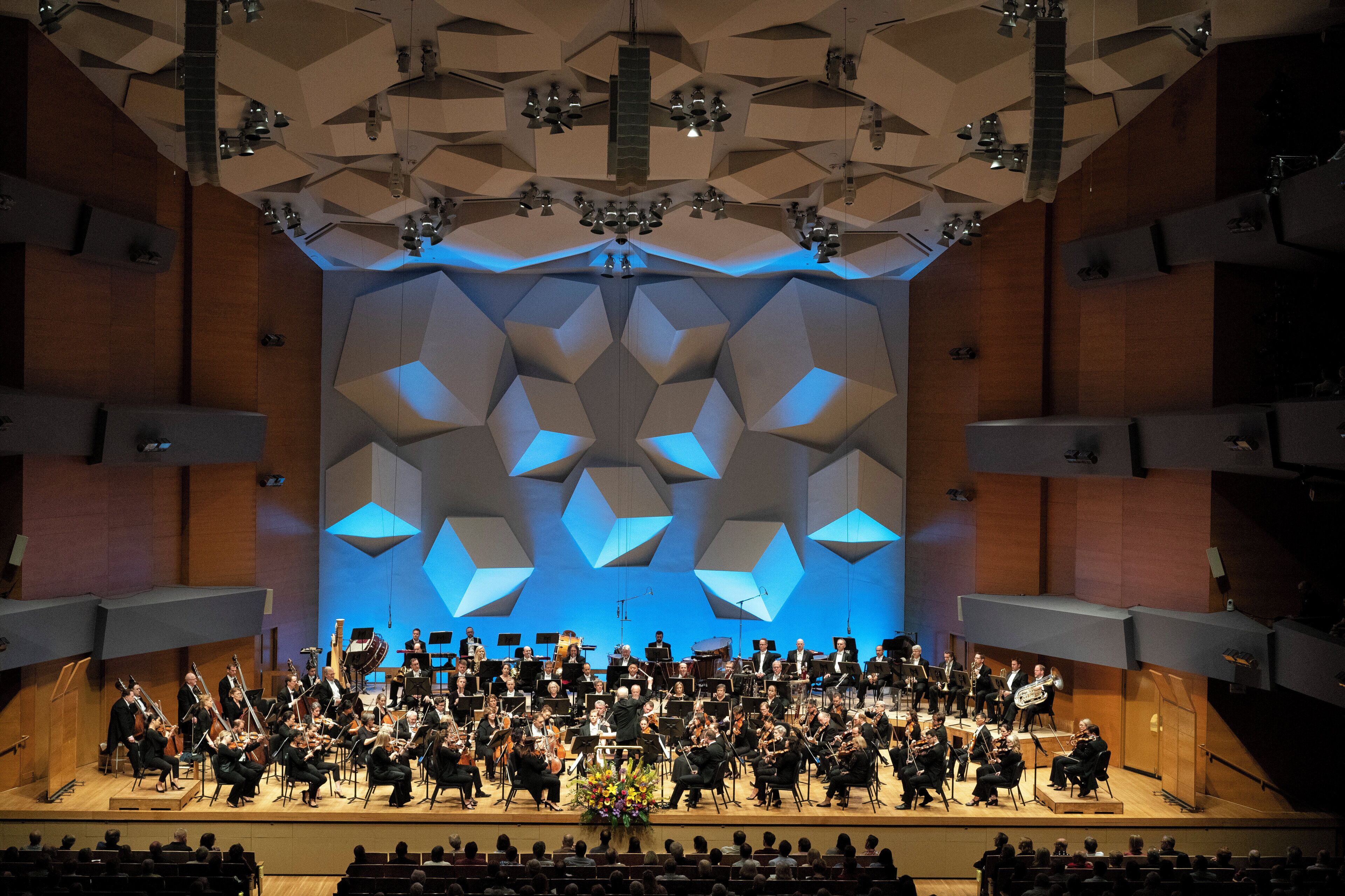 An orchestra performing on a stage with geometric ceiling design and seating for an audience.
