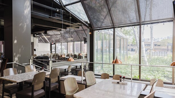Un restaurant moderne avec de hauts plafonds, de grandes fenêtres et une vue sur la ville. Les tables et les chaises sont disposées pour accueillir les clients.