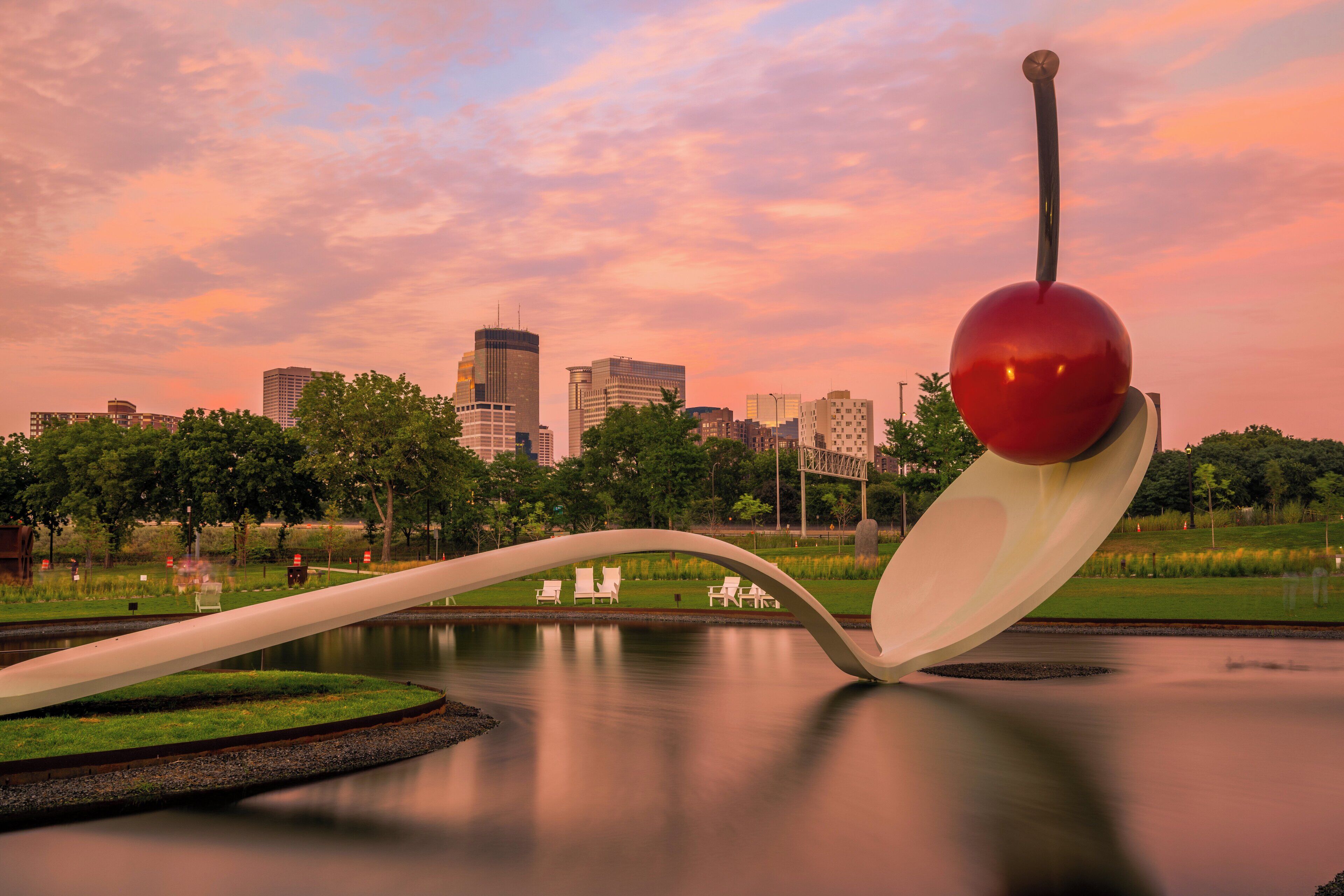 A park with a unique sculpture, a body of water, and a city skyline in the background.