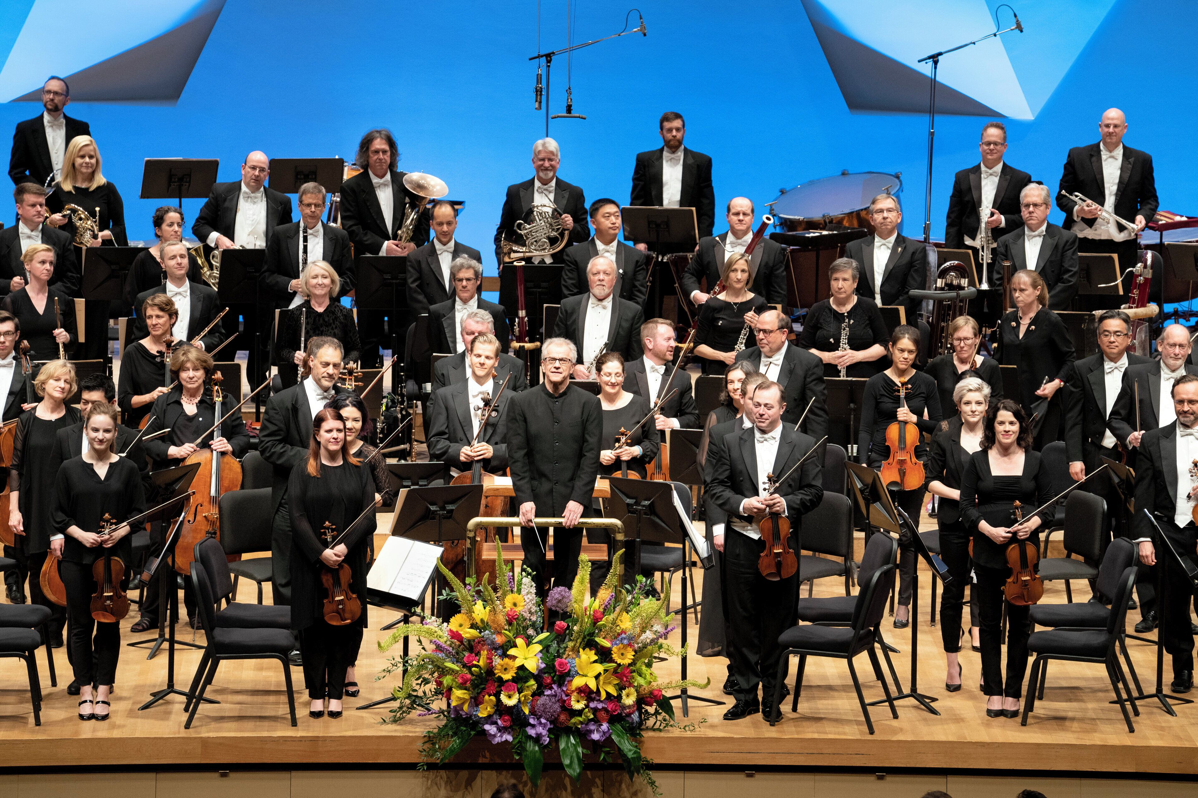 Un ensemble de musiciens, habillés de manière élégante, se produit sur scène, des chaises et une composition florale étant disposées autour d’eux.