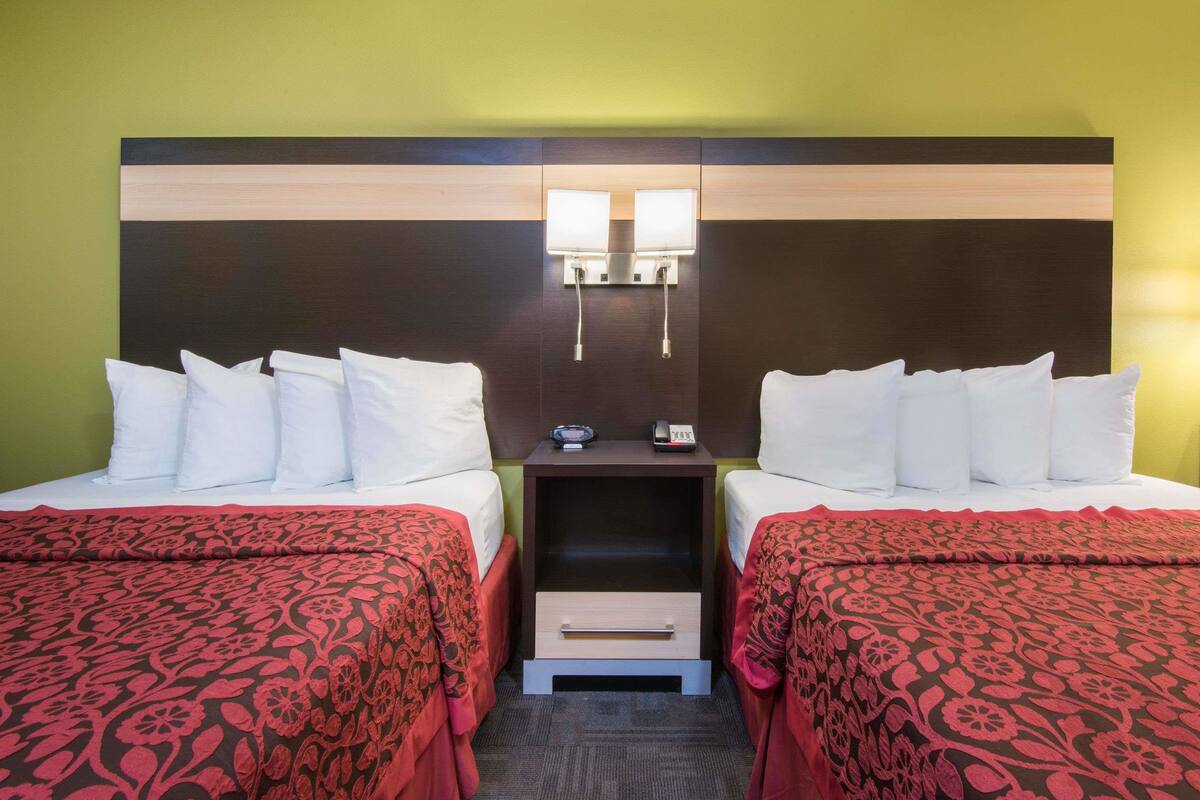 A hotel room with two beds, each with white linens and red patterned bedspreads. There is a nightstand between the beds with two wall-mounted lamps and a small drawer.