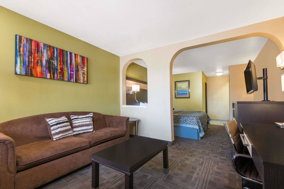 A hotel room with a brown sofa, a small table, a bed with a blue and white patterned bedspread, a TV mounted on the wall, and a colorful abstract painting on the wall.