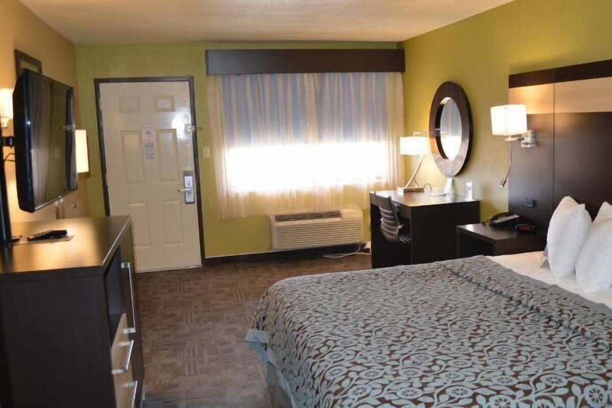 A hotel room with a bed, a desk, a chair, a mirror, a lamp, and a door.