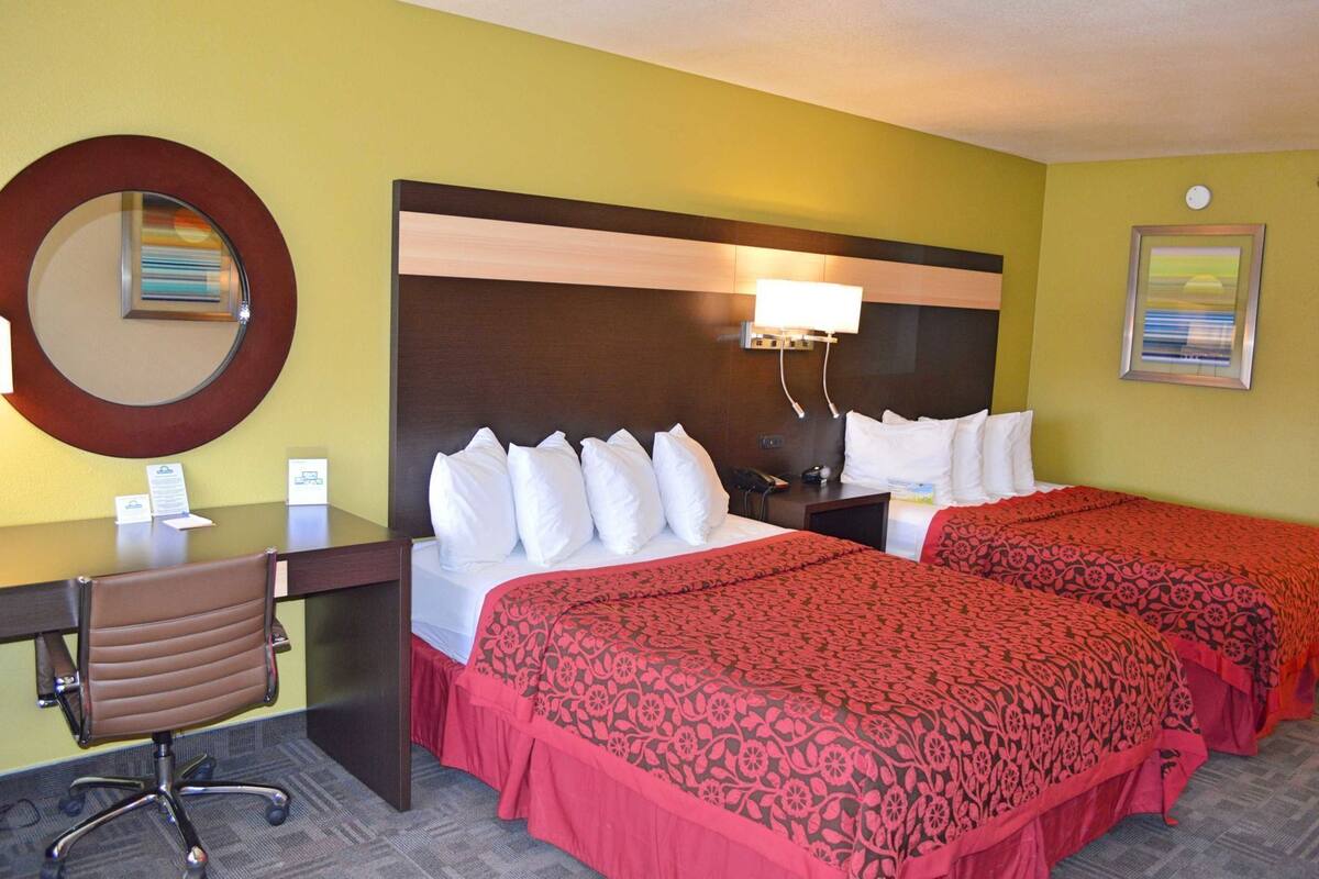A hotel room with two beds, a desk with a chair, a round mirror, a lamp, and a clock on the wall.
