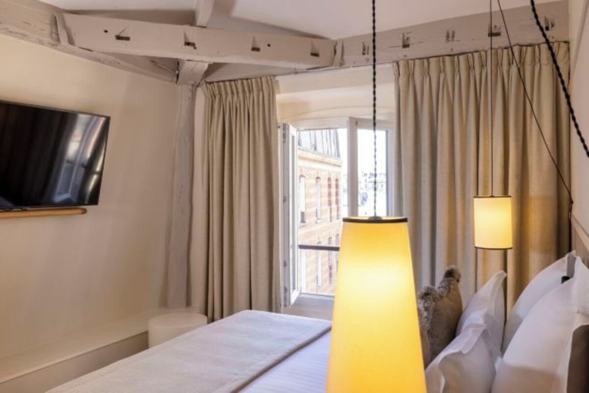 A hotel room with a bed, a TV mounted on the wall, a large window with curtains, and a hanging lamp.