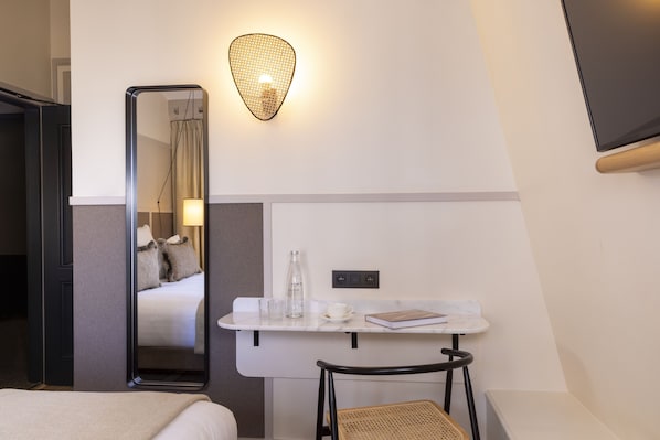 A hotel room with a bed, a desk with a mirror, a lamp, a bottle of water, a cup, and a book.