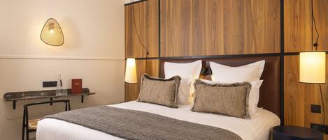 A hotel room with a large bed, wooden headboard, two bedside lamps, a small table with a chair, and a wall-mounted light fixture.