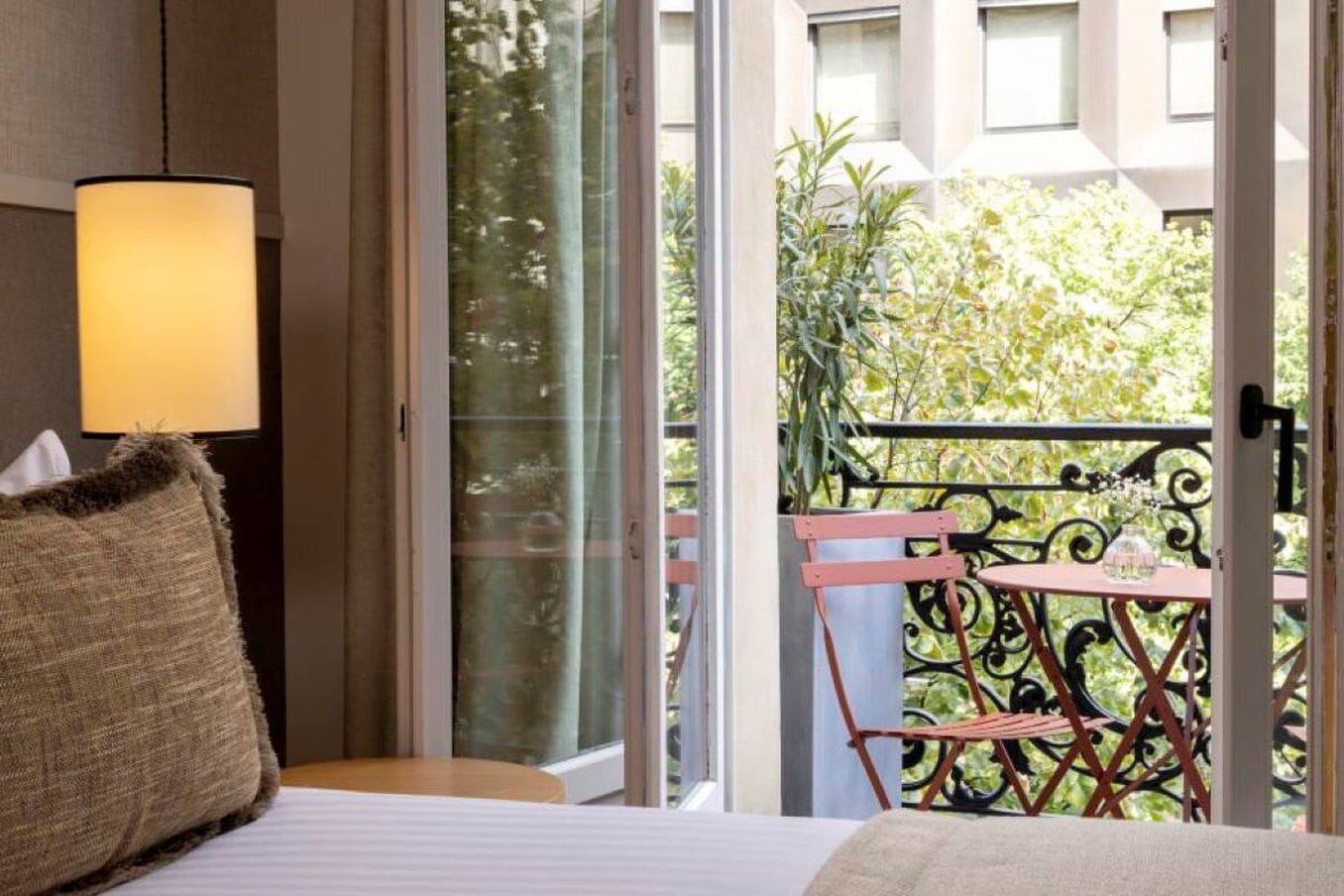 A hotel room with a bed, a bedside lamp, a window with a view of greenery, and a balcony with a chair and railing.