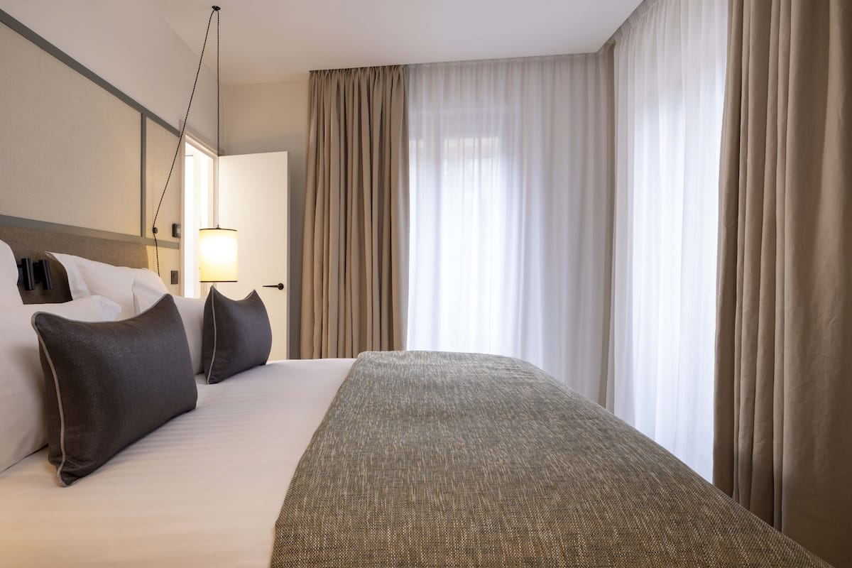 Ett hotellrum med en stor säng, en sänglampa och utsikt över omgivningarna genom tunna gardiner.