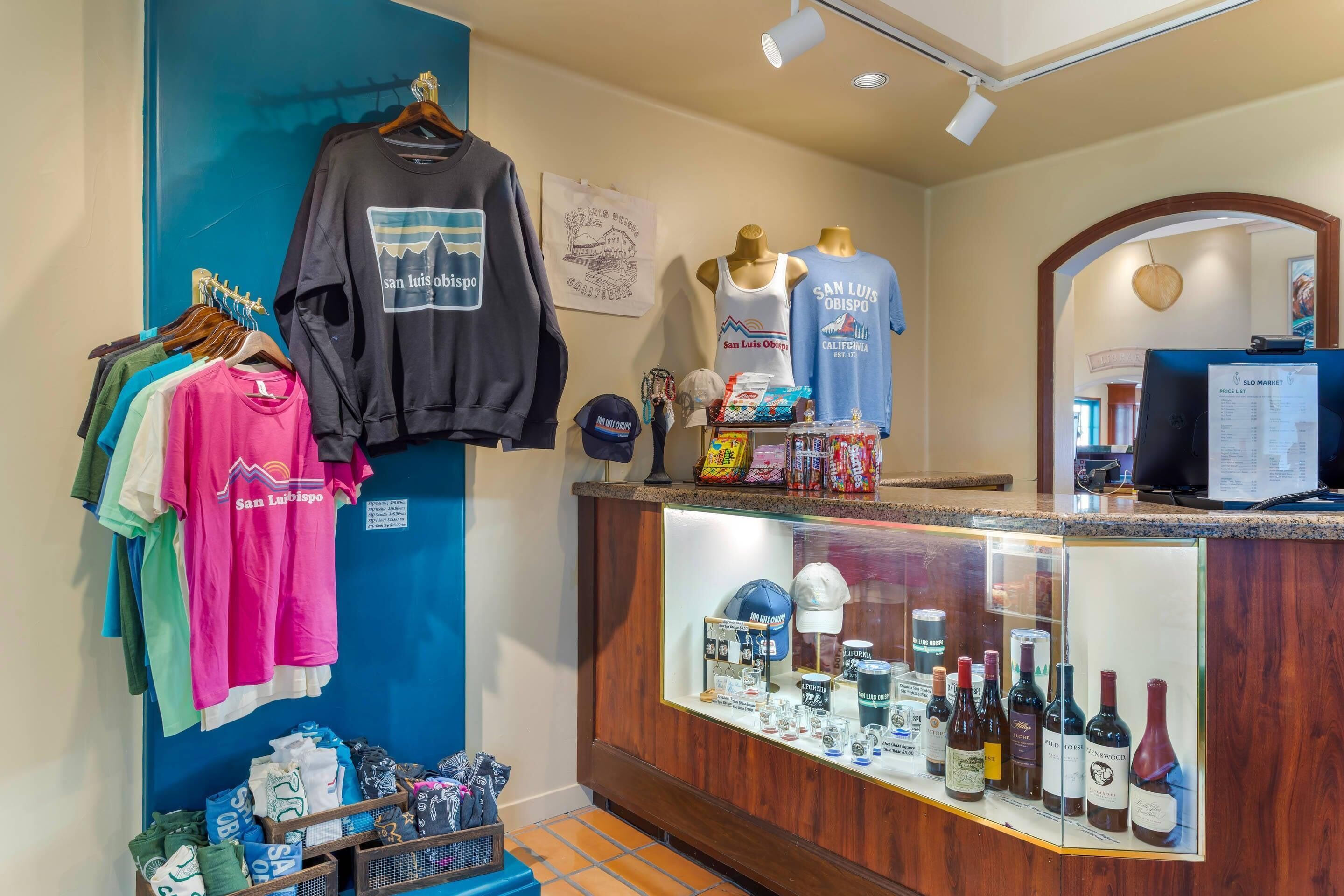 A retail space with clothing racks, a display of San Luis Obispo-themed apparel, and a wine selection behind a wooden counter.