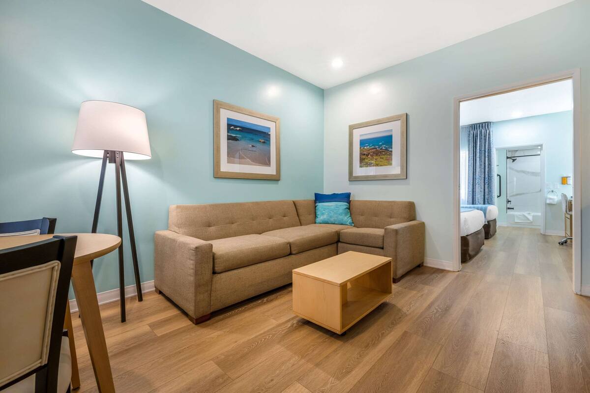 A modern living room with a sofa, coffee table, and floor lamp. There are framed pictures on the wall and a view into the bedroom.