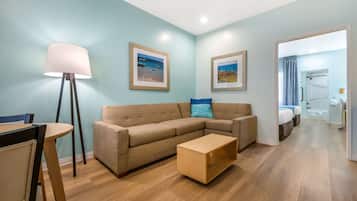 Un salon moderne avec un canapé, une table basse et une lampe sur pied. Des tableaux encadrés ornent le mur, et on aperçoit la chambre à coucher.