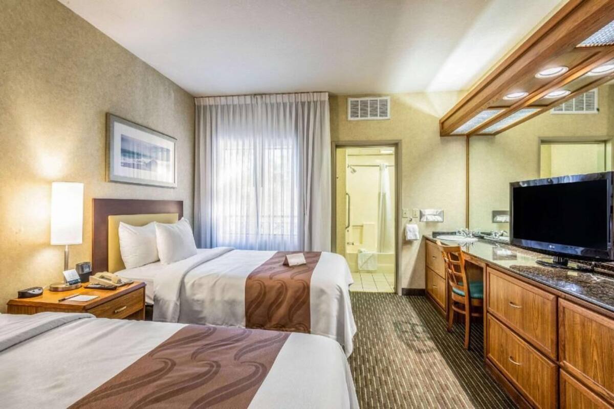 A hotel room with two beds, a TV, a desk with a chair, a bathroom visible through a door, and a large mirror.