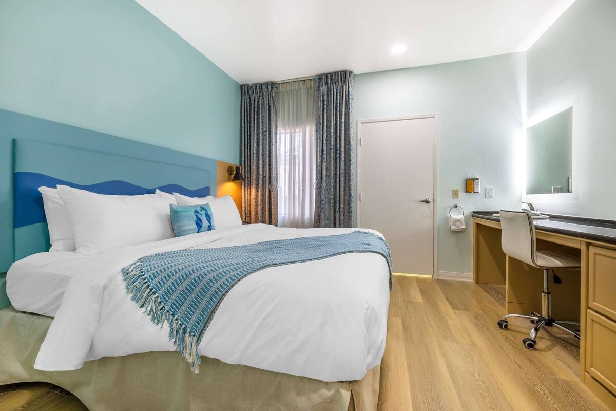 A hotel room with a bed, a desk, a chair, a mirror, and a door.