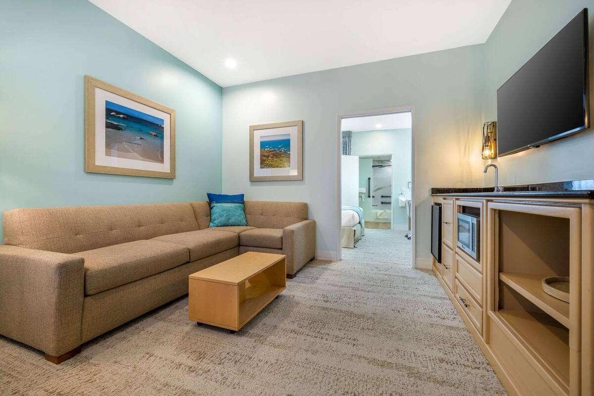 A living room with a beige sectional sofa, a wooden coffee table, and a flat-screen TV mounted on the wall. There are framed pictures on the wall, and a hallway leading to a bathroom.