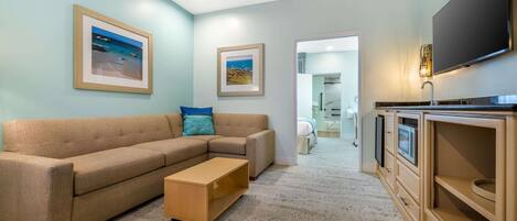 A living room with a beige sectional sofa, a wooden coffee table, and a flat-screen TV mounted on the wall. There are framed pictures on the wall, and a hallway leading to a bathroom.