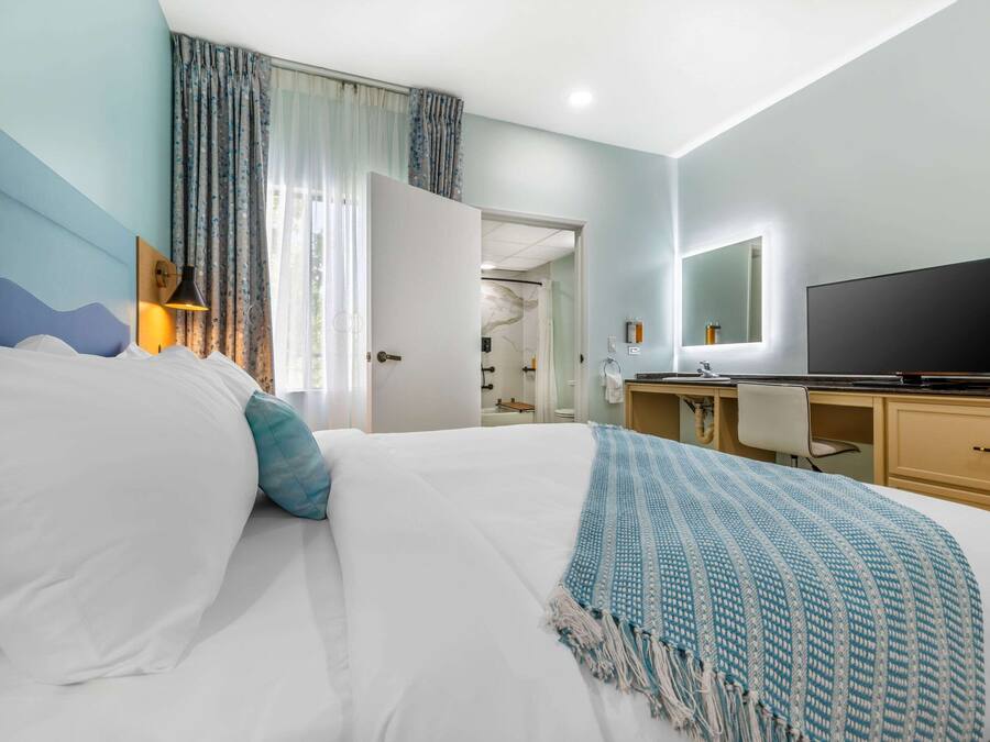 A hotel room with a bed, a desk, a TV, and a bathroom visible through an open door.