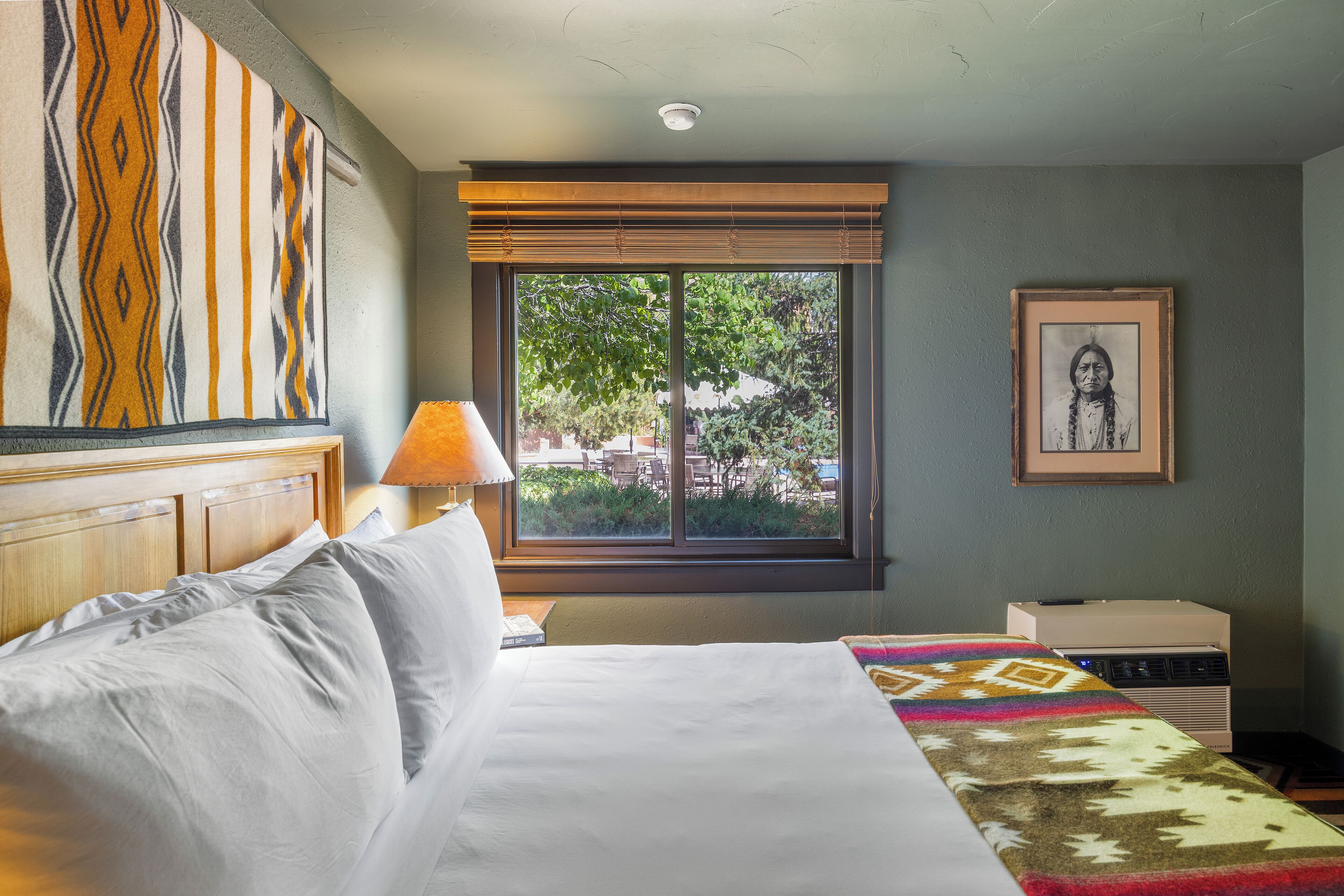 A hotel room with a bed, a window with blinds, a lamp, a framed picture on the wall, and a small table with a box.
