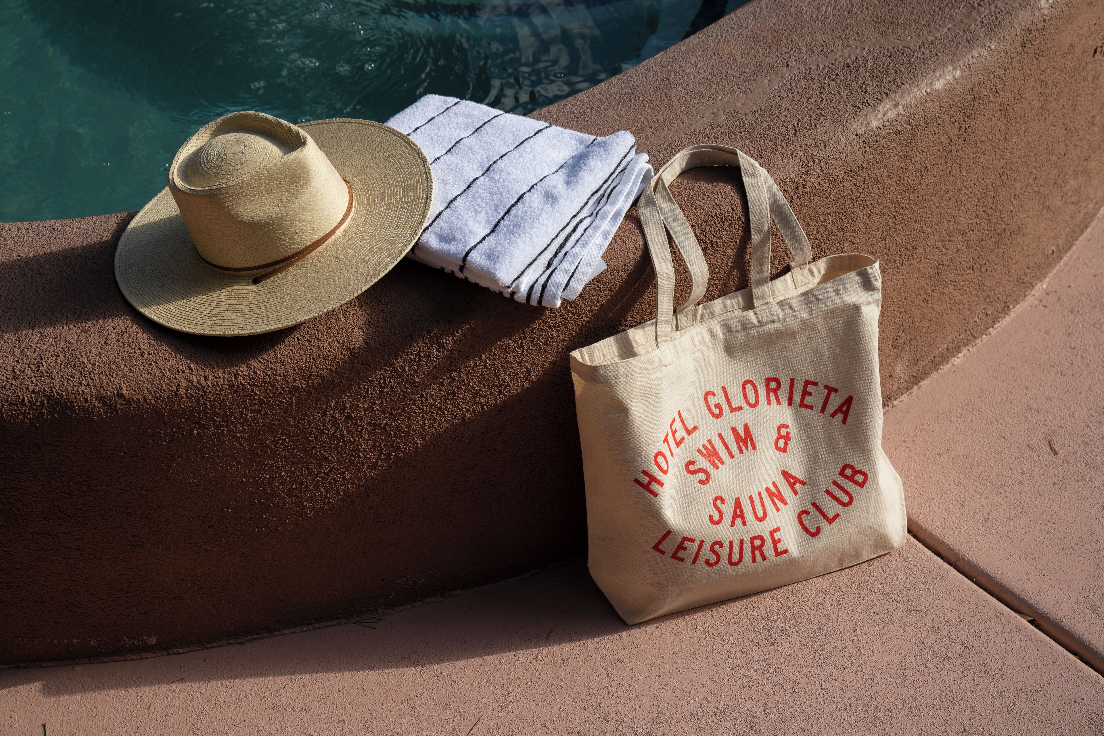 A straw hat, a striped towel, and a tote bag with text on a poolside surface.