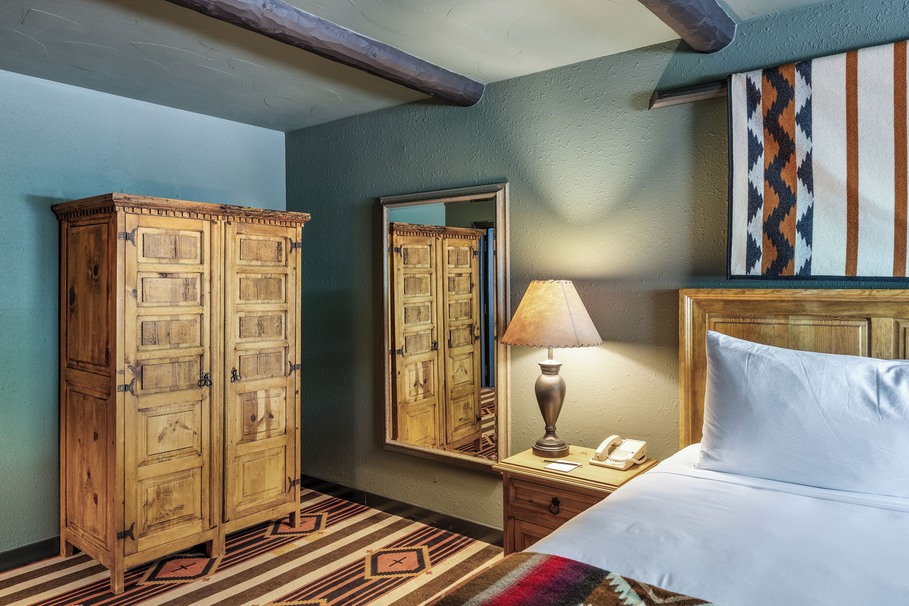 A hotel room with a large wooden wardrobe, a bedside table with a lamp, and a bed with white linens.