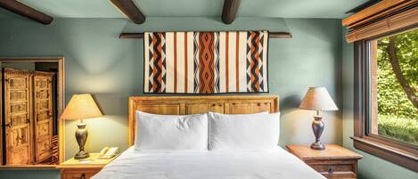 A hotel room with a large bed, wooden headboard, a patterned wall hanging, a window with a view of greenery, and a traditional wooden door.
